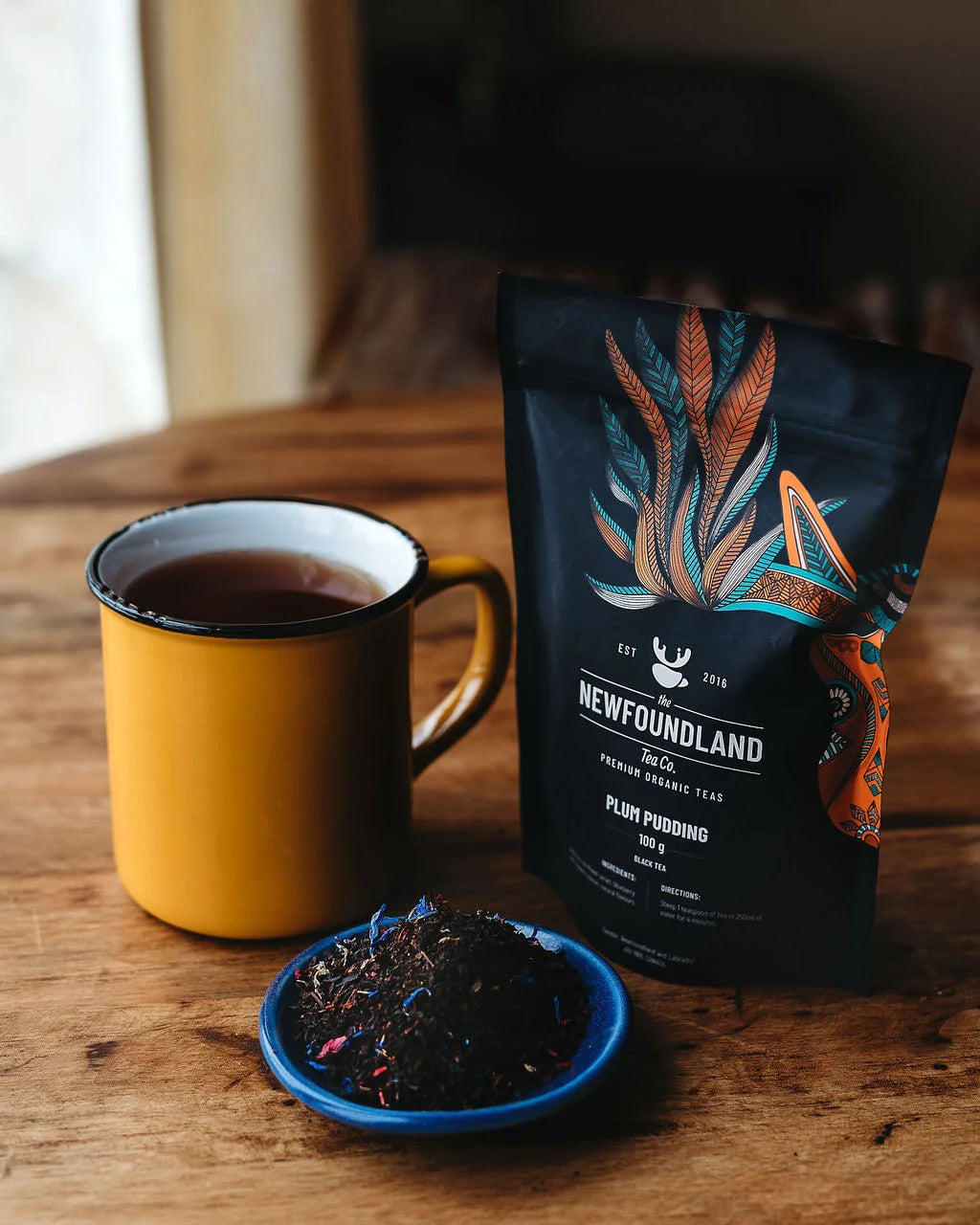 Plum Pudding black tea pouch beside a yellow enamel mug and loose tea with cornflower petals