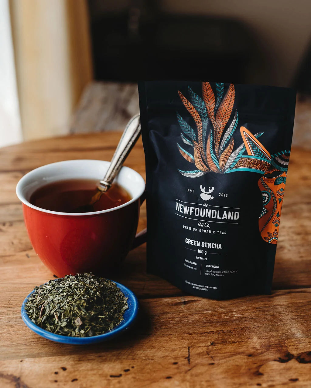 Sencha green tea pouch with a red cup of tea and a dish of loose sencha leaves